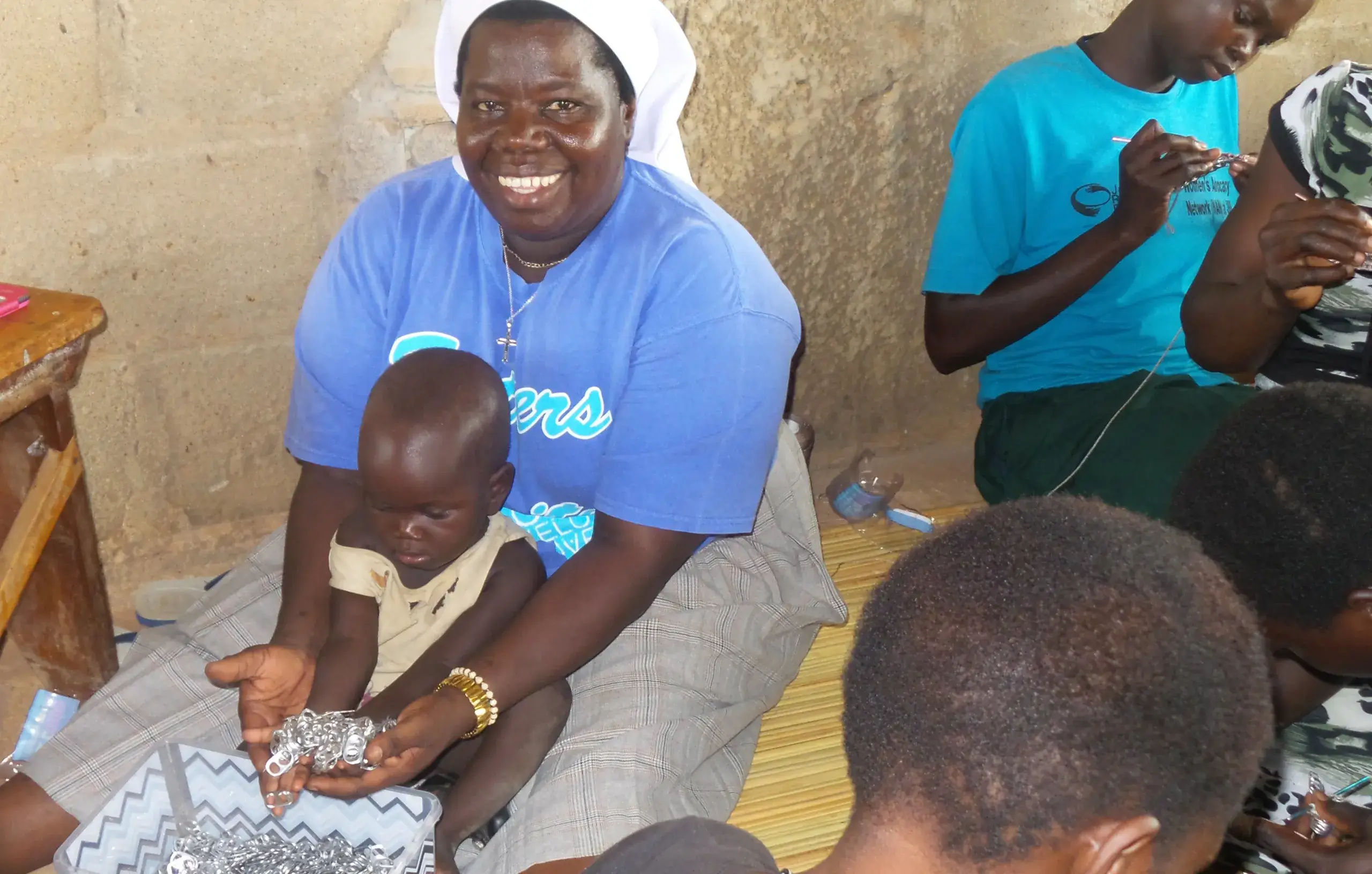 Sister Rosemary holding child near women sewing pop tab purses
